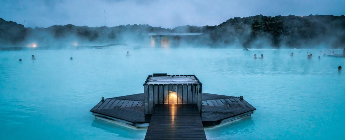 Geothermal lagoon in Iceland with a wooden dock, surrounded by mountains. People are swimming in the milky blue water.