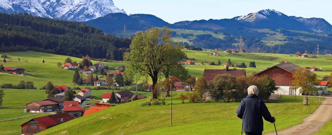 A person with walking poles on a path in the green, rural landscape of Riezlern, with small houses and snow-capped mountains in the background.